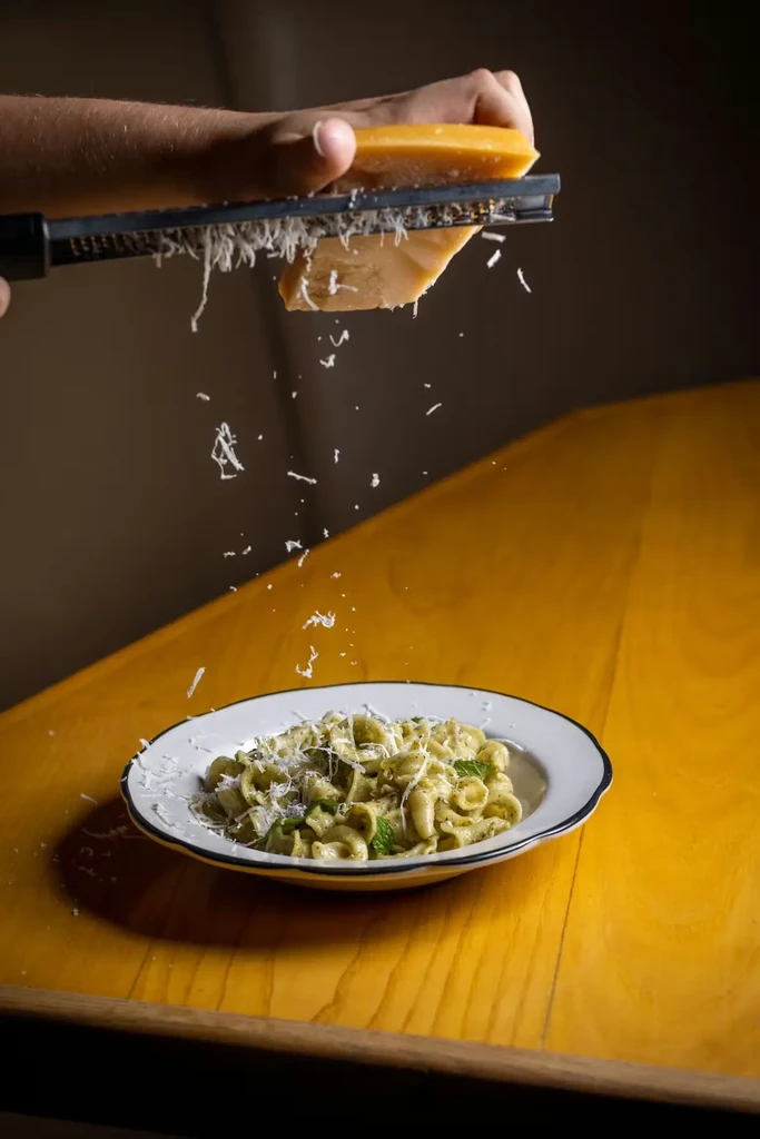 Hand grating parmesan cheese onto a bowl of green shell pasta.