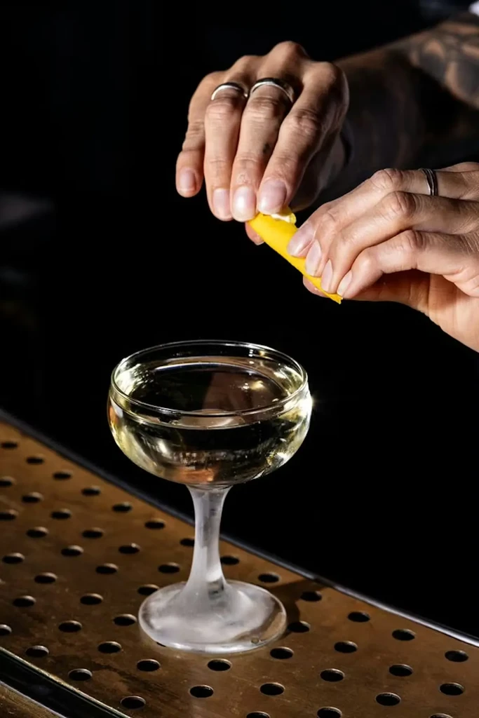 Hands squeeze citrus peel over a clear cocktail in a coupe glass.