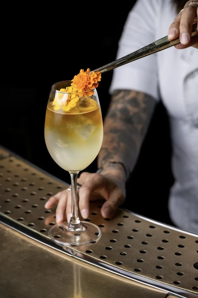 Tattooed hands garnish a two-toned cocktail with an orange marigold flower.