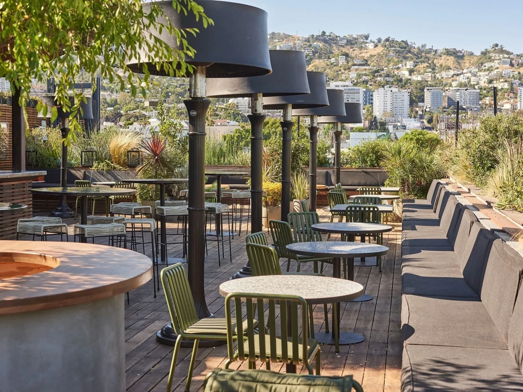 Sunlit rooftop dining patio with seating, heaters, plants, city view.