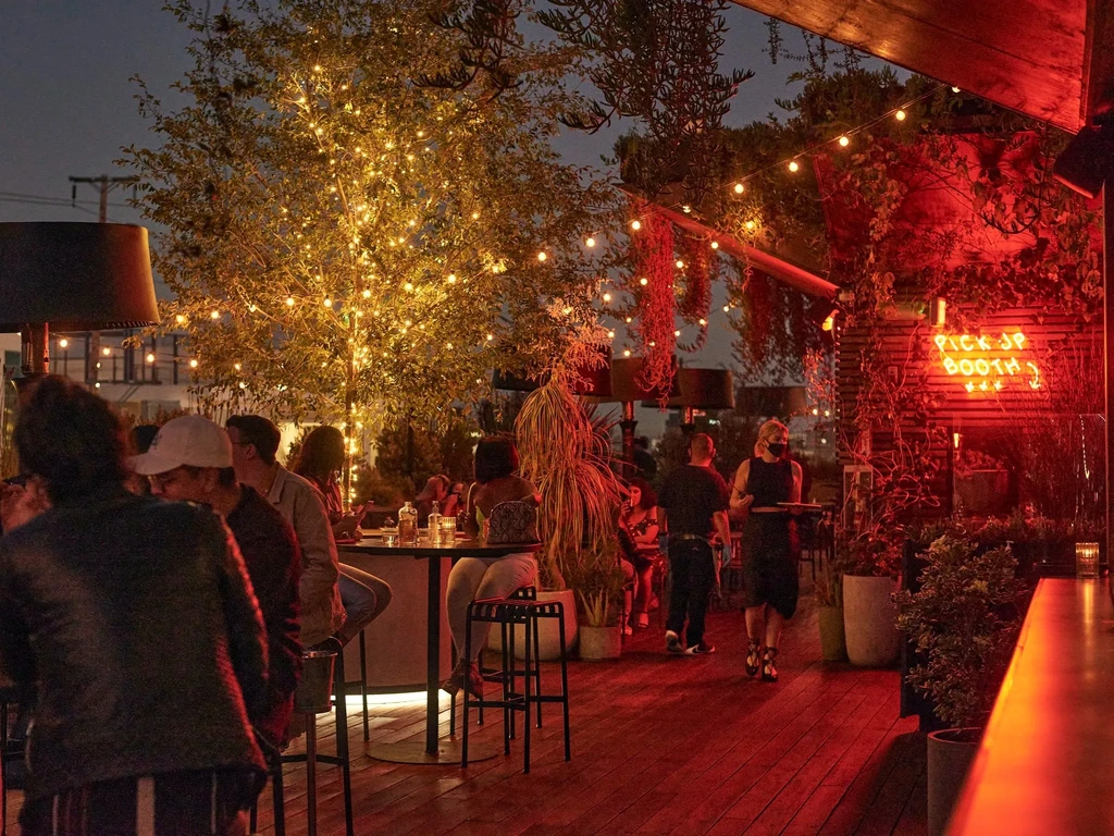 Nighttime outdoor restaurant with string-lit tree, neon sign, and patrons.