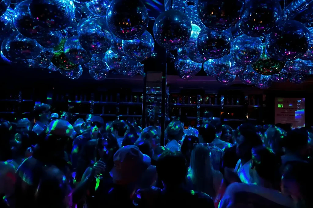 Disco balls cast blue-green shimmering light on a lively dancing crowd.