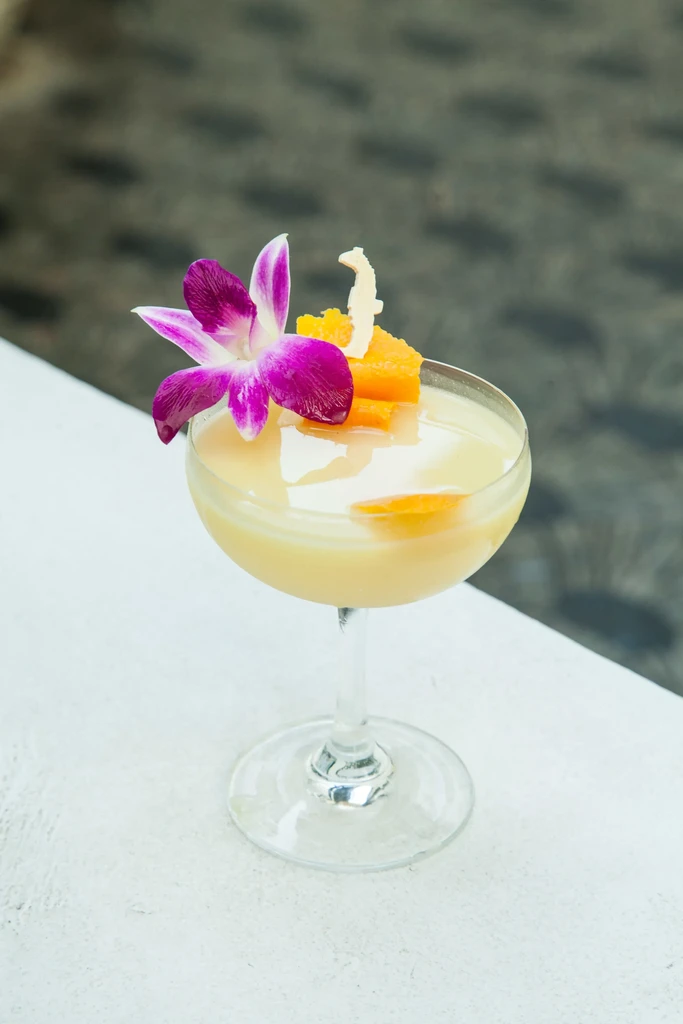 Pale yellow cocktail in coupe glass with purple orchid and seahorse garnish.