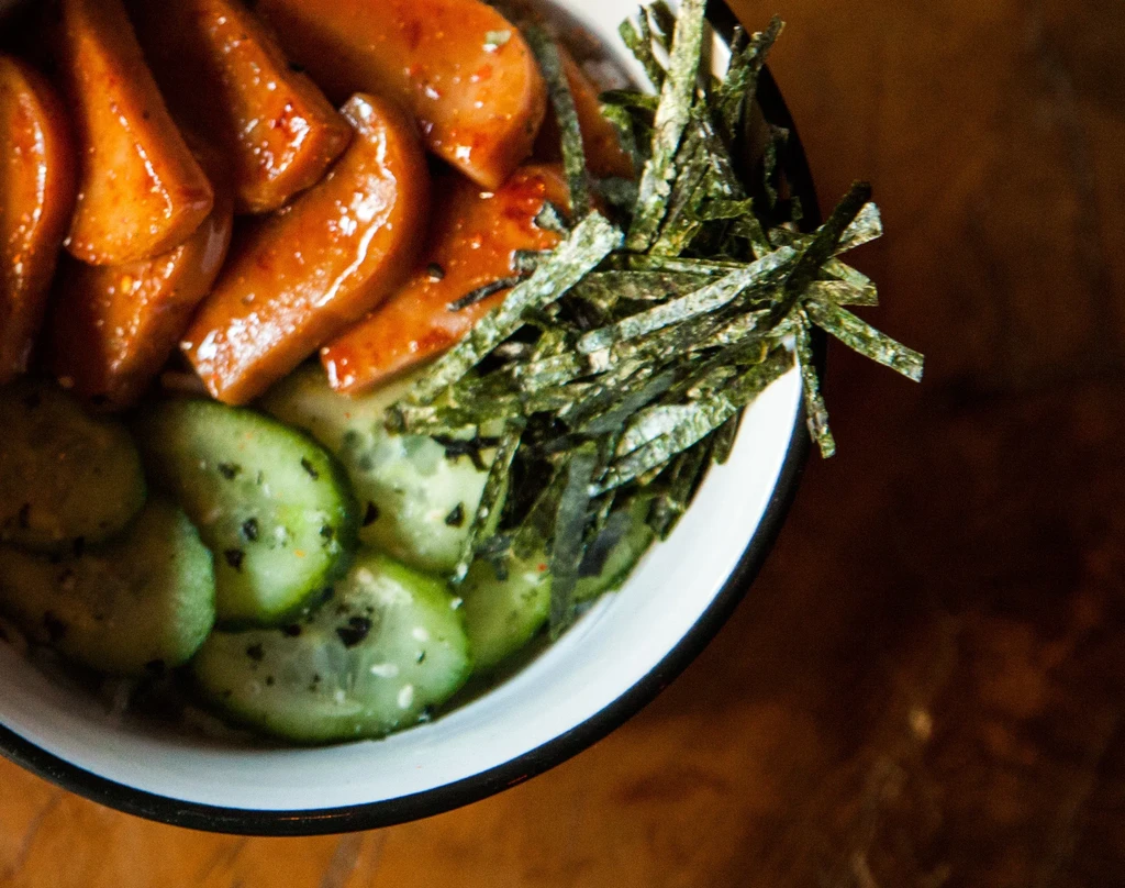 Close-up food bowl with glazed food, cucumber, and shredded nori.