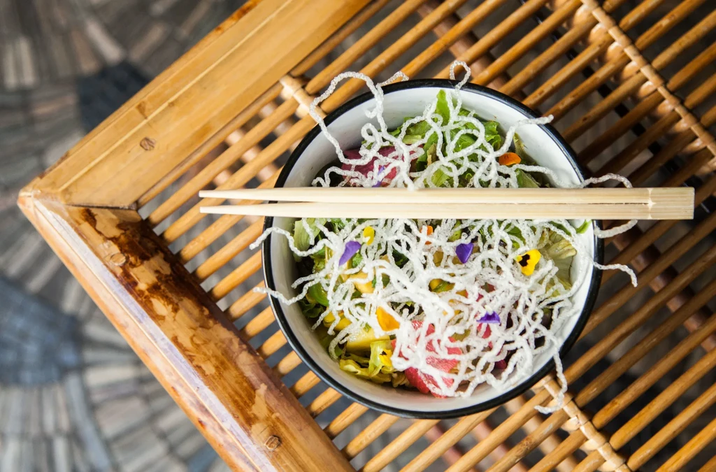 Colorful salad with crispy noodles, edible flowers, and chopsticks on bamboo.