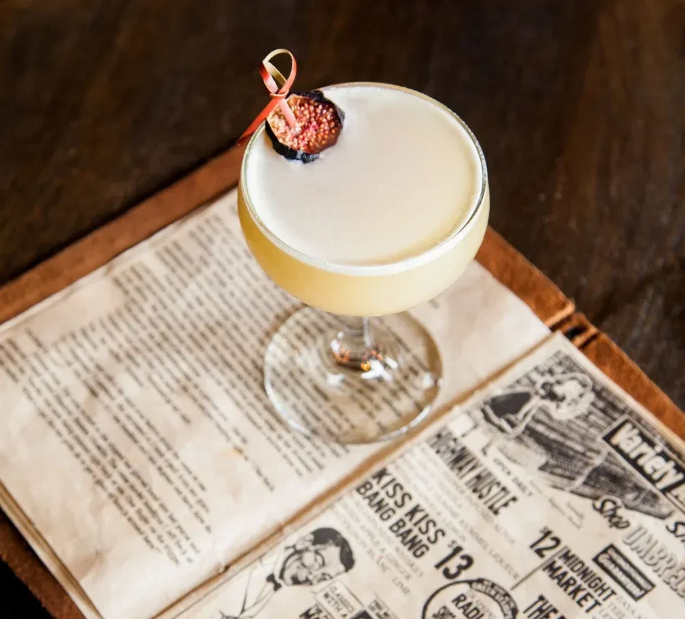 Light yellow cocktail with fig and ribbon garnish on a newspaper menu.