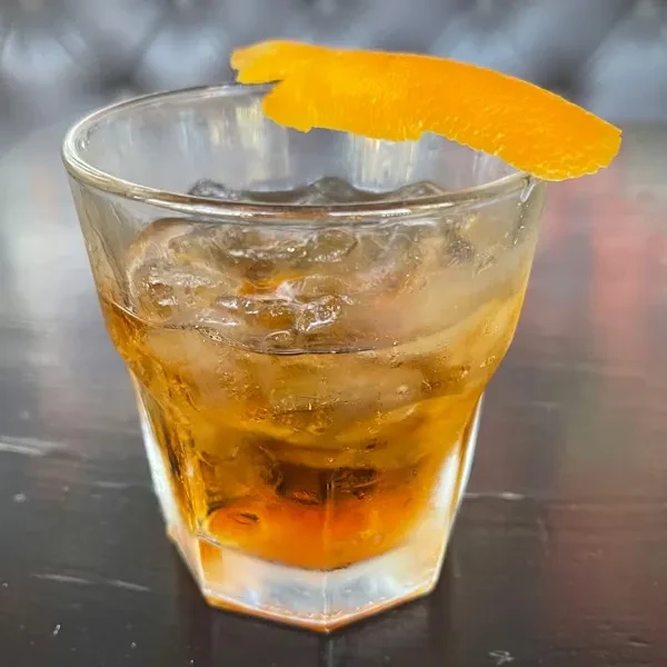 Dark amber cocktail in faceted glass with ice and orange peel garnish.