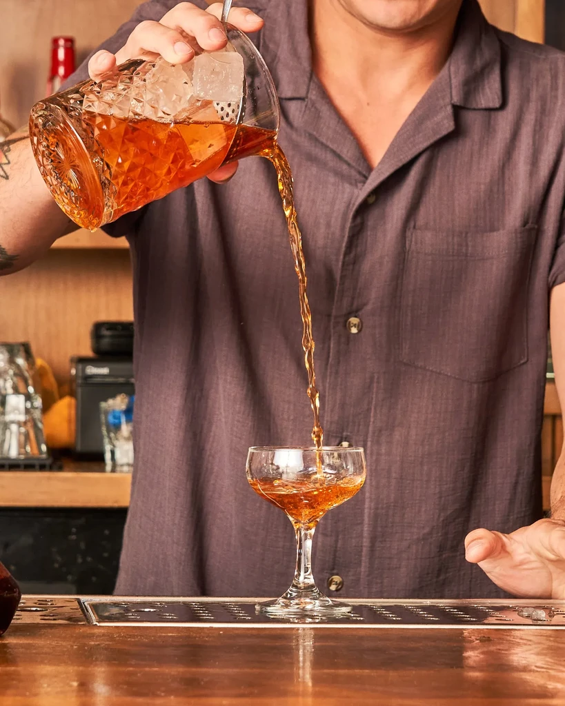 Bartender pours amber cocktail from mixing glass into coupe glass.