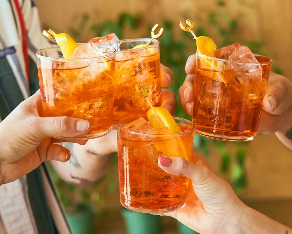 Four hands toast orange cocktails with ice, citrus, and decorative skewers.