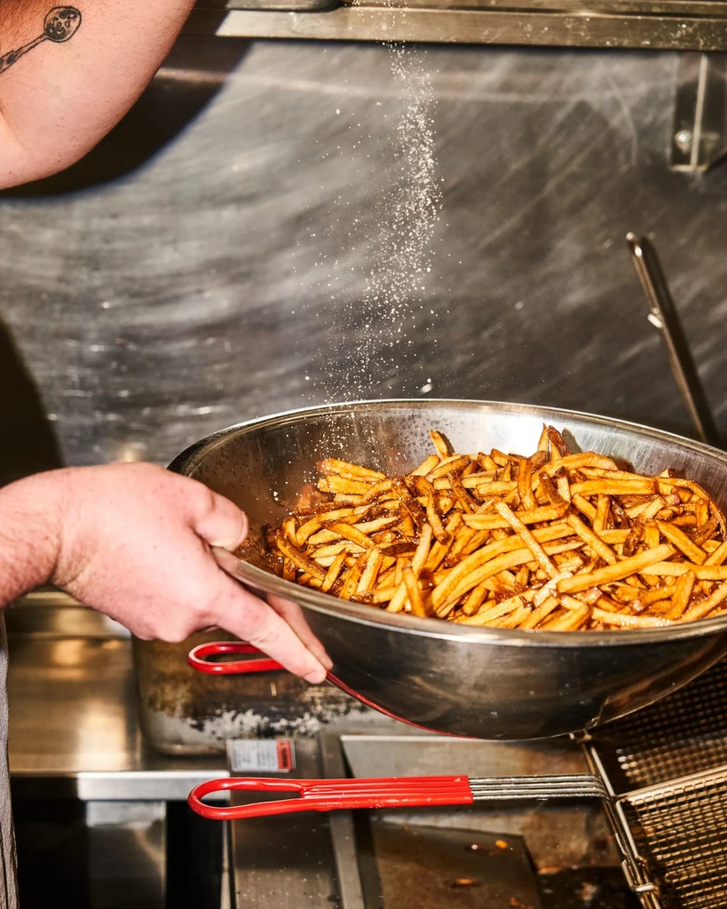 Close-up of golden french fries being salted in a metal bowl.