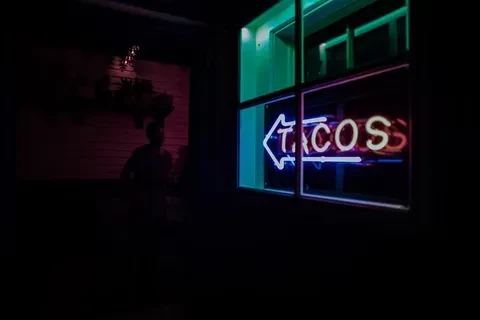 Bright neon "TACOS" sign with blue arrow in dark restaurant window.