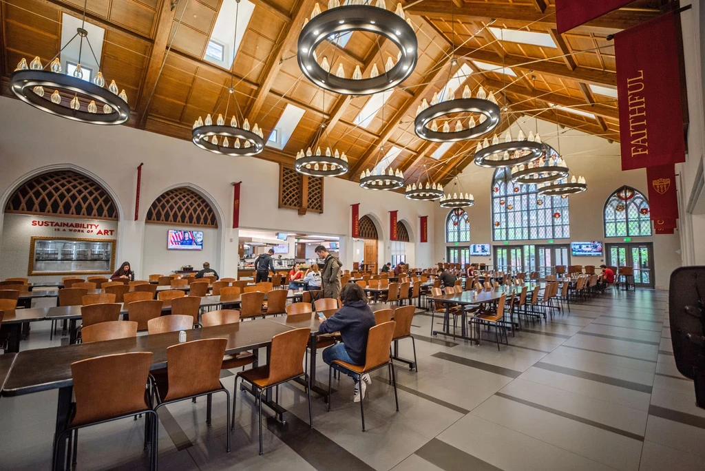Grand dining hall with wooden beamed ceiling, chandeliers, and people.