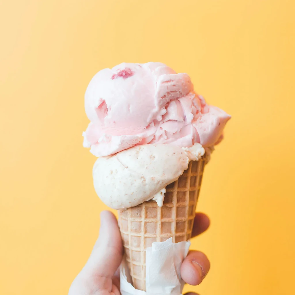 Hand holding a waffle cone with pink and cream ice cream on yellow background.