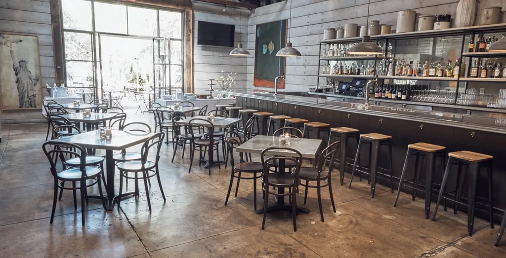 Modern rustic restaurant interior with dining tables, long bar, and patio view.