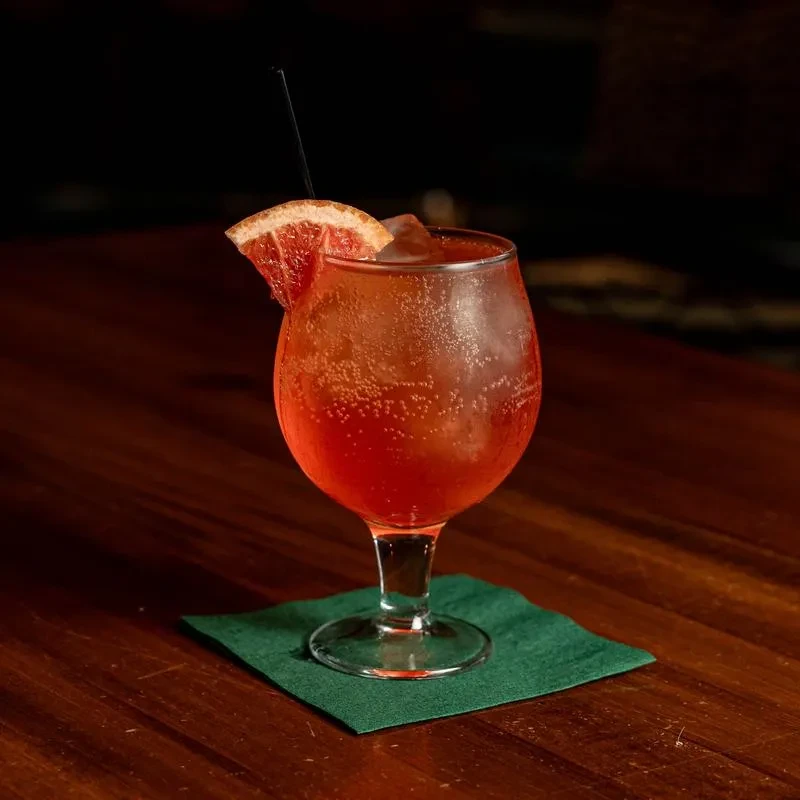 Carbonated red-orange cocktail with ice, grapefruit, and straw on a table.