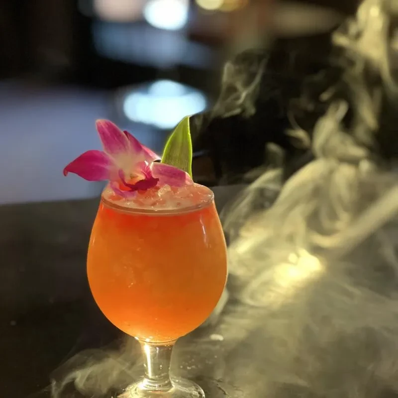 Stemmed orange cocktail, pink orchid, green leaf, and swirling smoke.