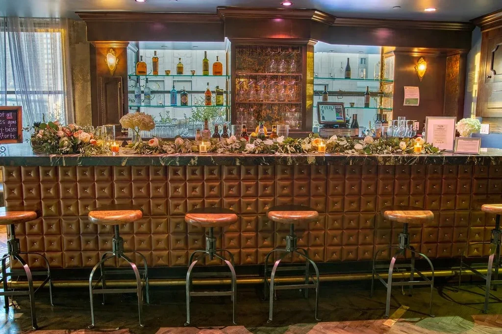 Tufted brown bar, green marble top, floral garlands, liquor bottles, barstools.