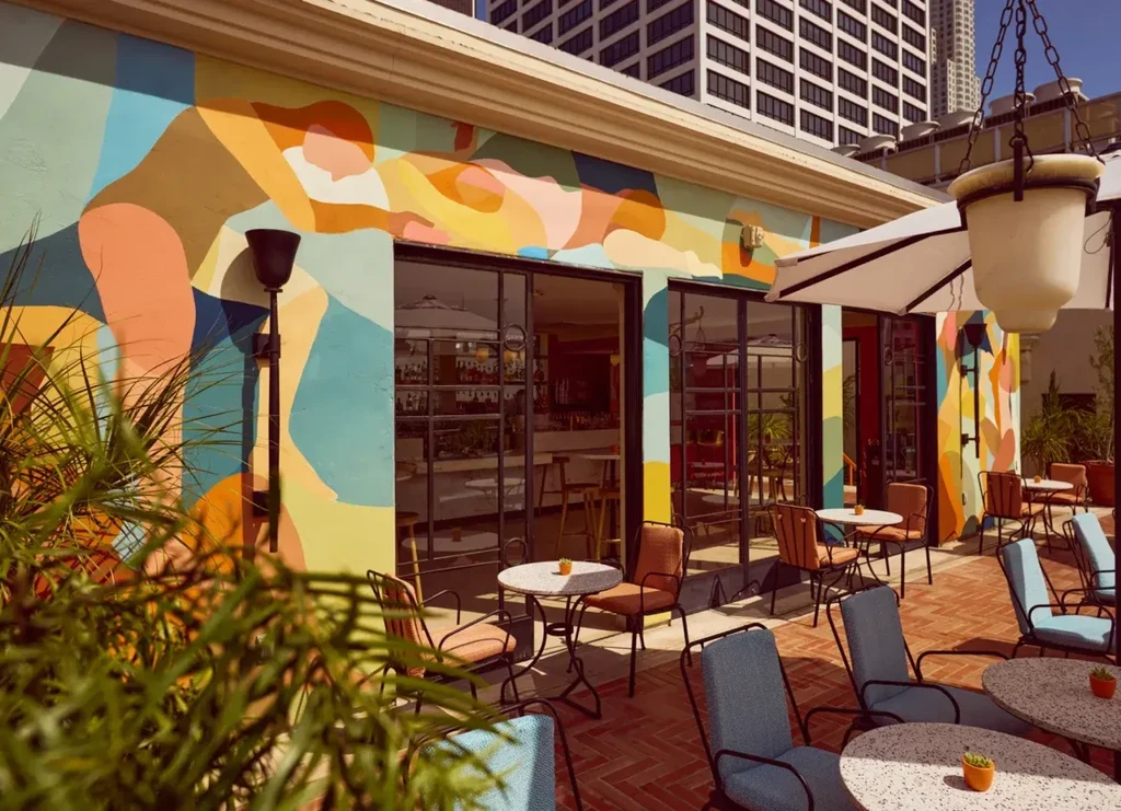 Restaurant patio with colorful abstract mural, tables, and urban buildings.