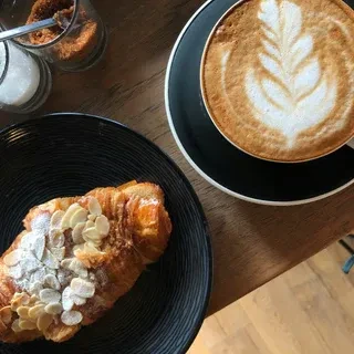 Overhead: Golden almond croissant, latte with leaf art, sugar, wooden table.