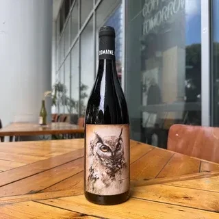 Red wine bottle with stylized owl face label on wooden table.