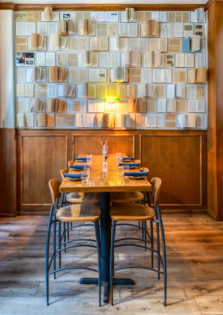 Warm restaurant interior with wooden table, stools, and book-themed wall art.