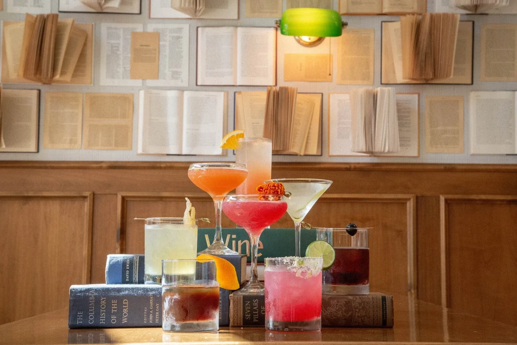 Seven colorful cocktails on old books, against a wall of open books.