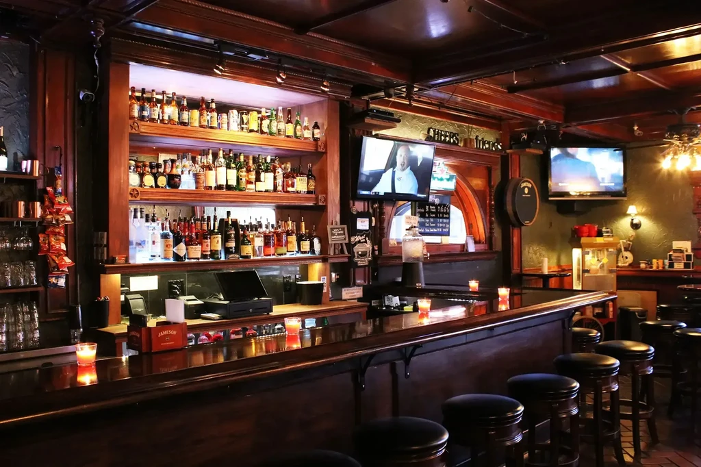 Dimly lit dark wood bar with backlit liquor bottles and two TV screens.