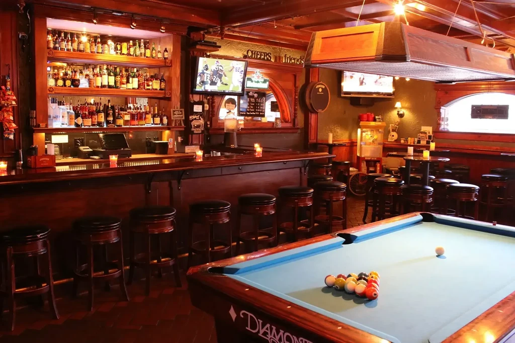 Warmly lit bar interior with stocked counter, blue pool table, and sports.