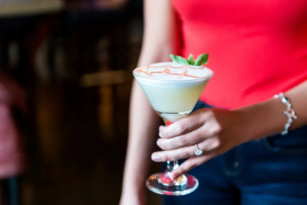 Hand holds pale yellow cocktail with frothy top, red swirl, mint garnish.
