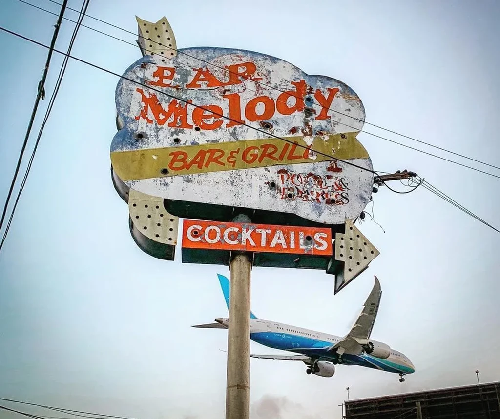Weathered 'Bar Melody' sign with airplane flying low under overcast sky.