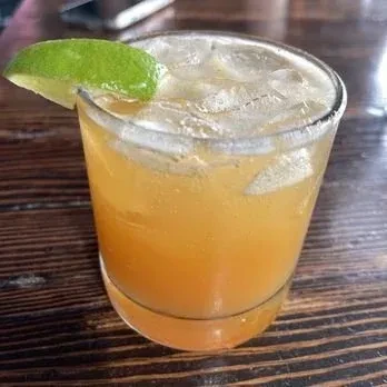 Amber drink with ice and lime wedge in glass on wooden table.