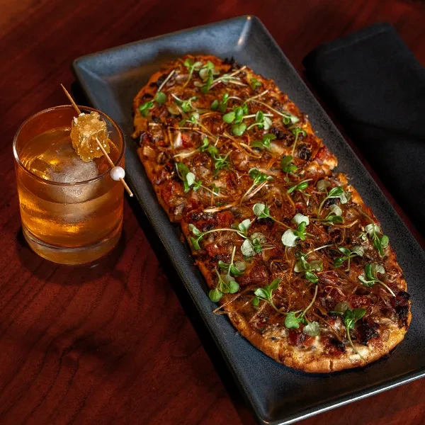 Gourmet flatbread with microgreens next to an amber cocktail with honeycomb.