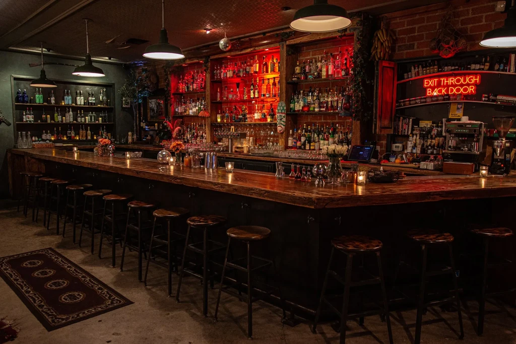 Dimly lit bar interior with rustic counter, stools, and glowing liquor shelves.
