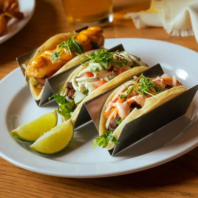 Three tacos in metal holders with various toppings and lime wedges.