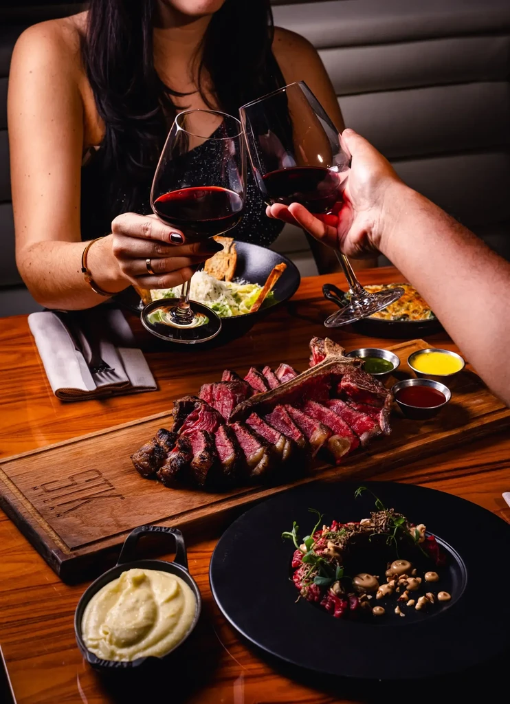 Two people toast red wine over a large, sliced steak.