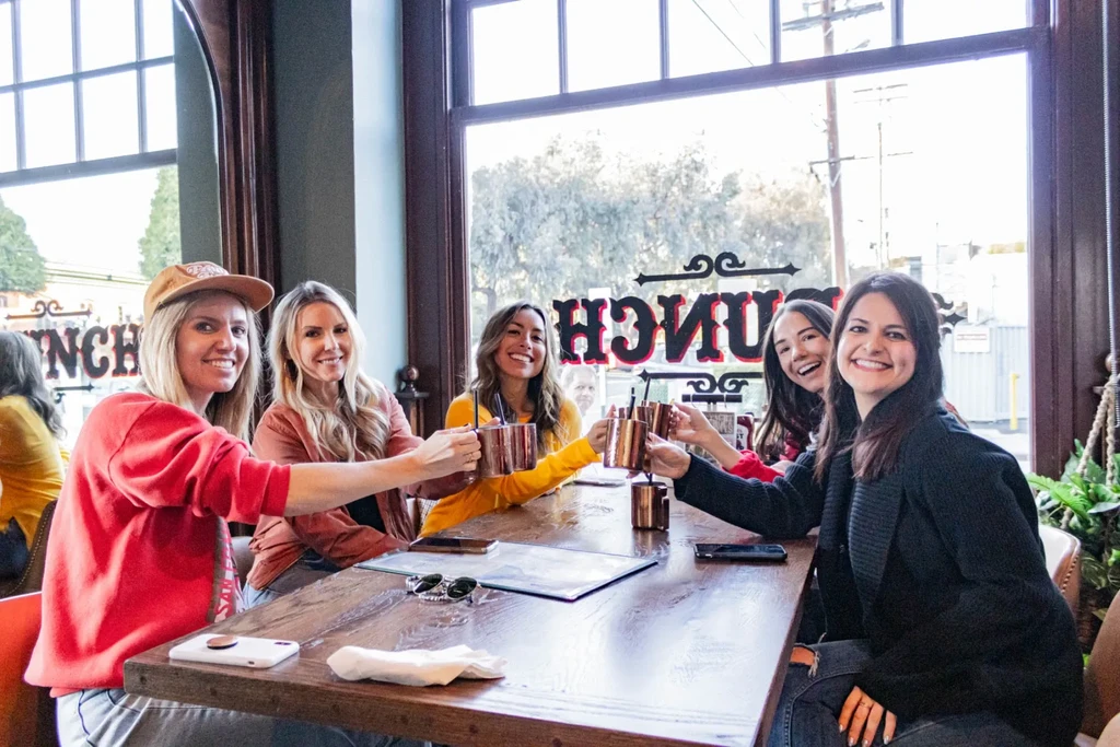 Five smiling women toast with copper mugs at a restaurant table.
