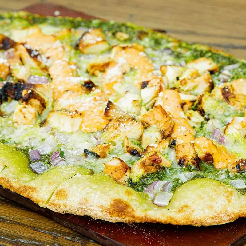 Rectangular pizza with green pesto, grilled chicken, red onion, cheese.