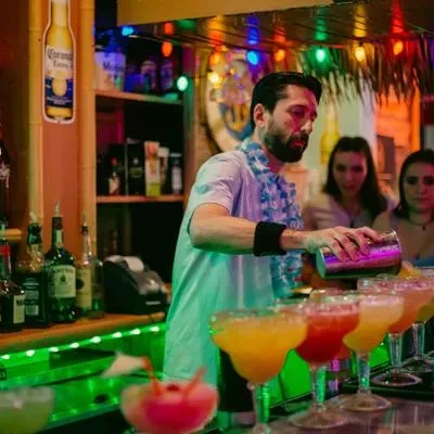 Bearded bartender in lei shakes colorful cocktails at green-lit tiki bar.