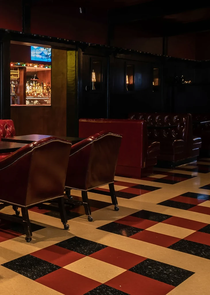 Dark restaurant interior with red leather chairs, checkerboard floor, and backlit bar.