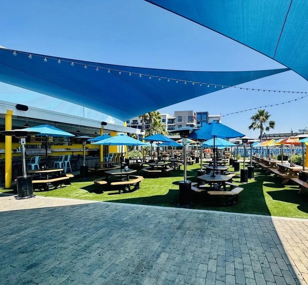 Vibrant outdoor beer garden with picnic tables, yellow bar, blue shade.