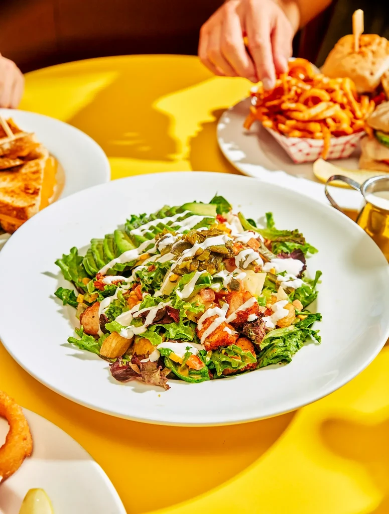 Colorful salad, curly fries, and mini burgers on a yellow table.