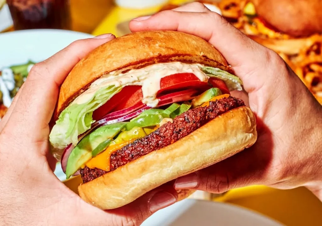 Vibrant gourmet burger with avocado, cheese, and vegetables held by hands.