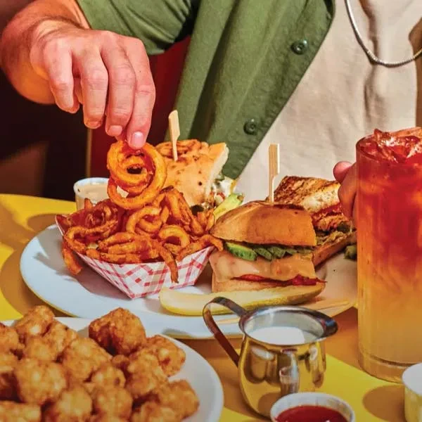 Hand picks curly fry from casual meal spread on yellow table.
