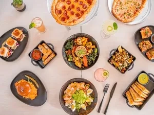 Overhead flat lay of diverse pizzas, salads, sandwiches, and cocktails.