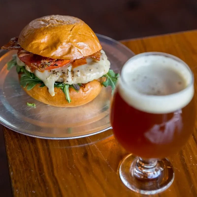 Grilled burger with melted cheese, bacon, and amber beer on wood.