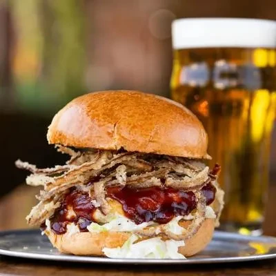 Close-up: large burger with fried onions, sauces, lettuce, and a beer.