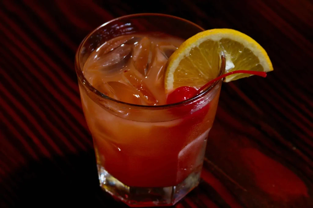 Close-up of a layered orange-red cocktail with ice, orange, and cherry.