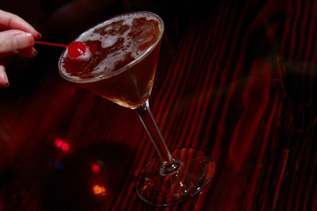 Amber cocktail in martini glass with hand adjusting a red cherry.