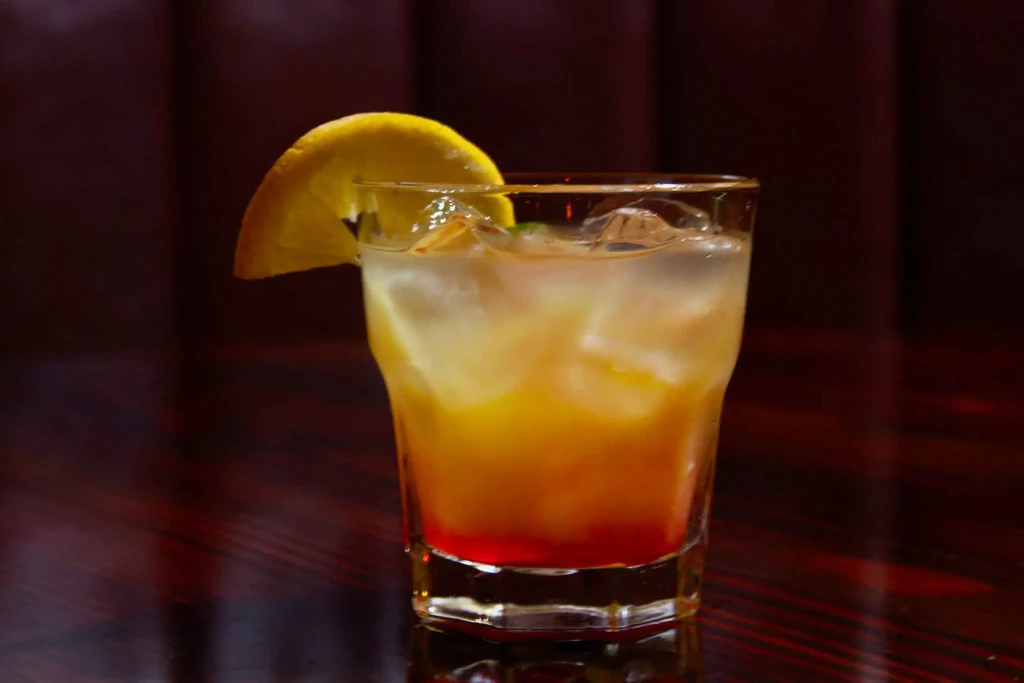 Layered cocktail with red, orange, yellow gradient, ice, and orange garnish.