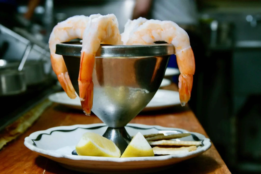 Shrimp cocktail in silver bowl, lemon, crackers on scalloped plate.